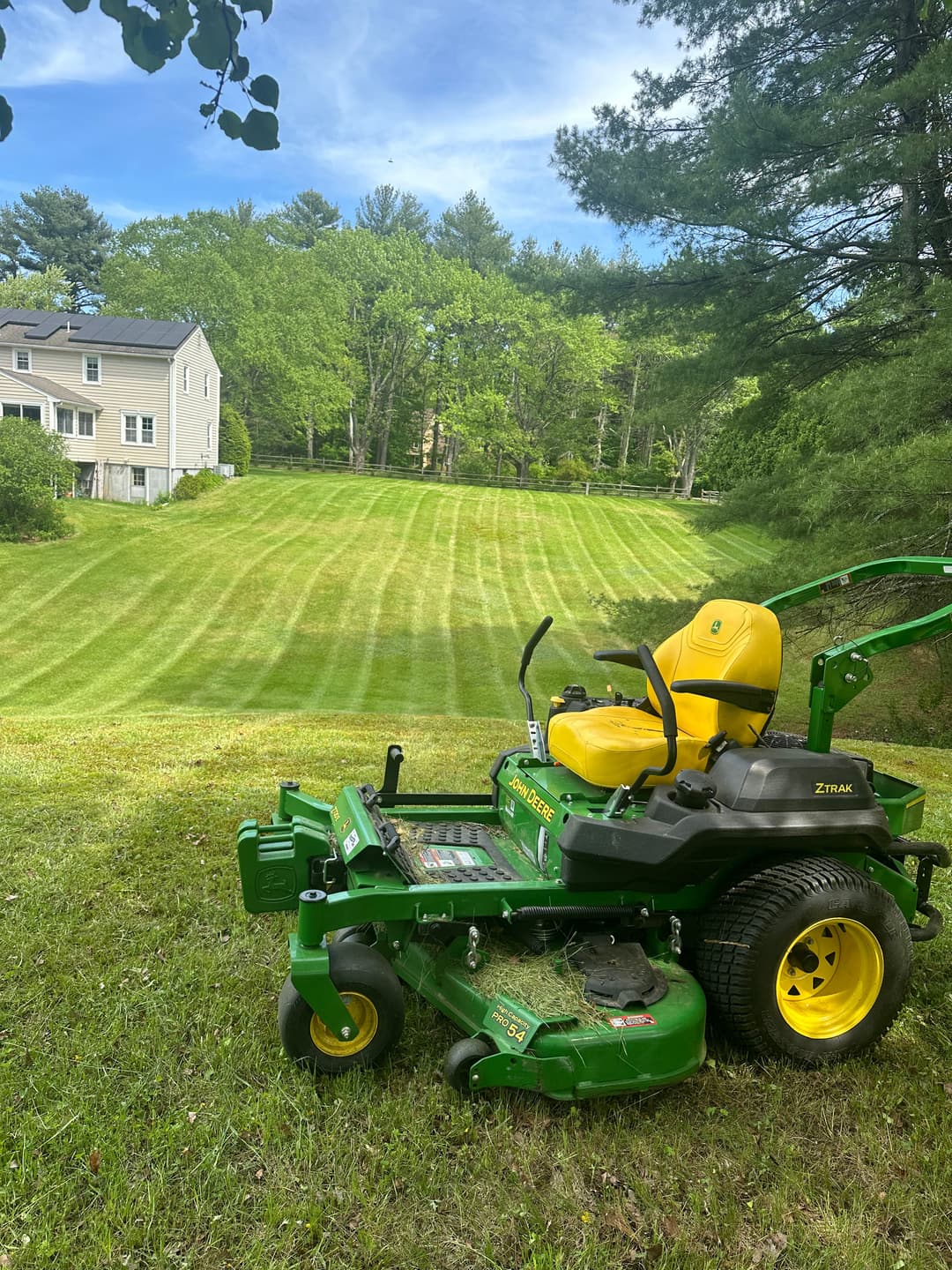 Green zero-turn lawn mower on a freshly cut lawn with trees and a house in the background.