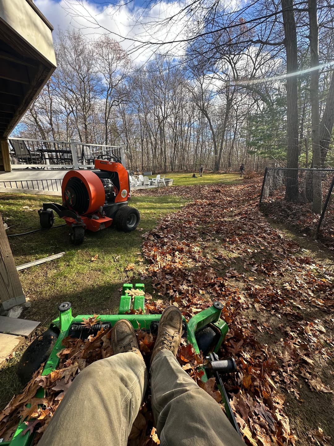 Person sitting on a riding lawn mower, surrounded by fallen leaves, with a leaf blower nearby.