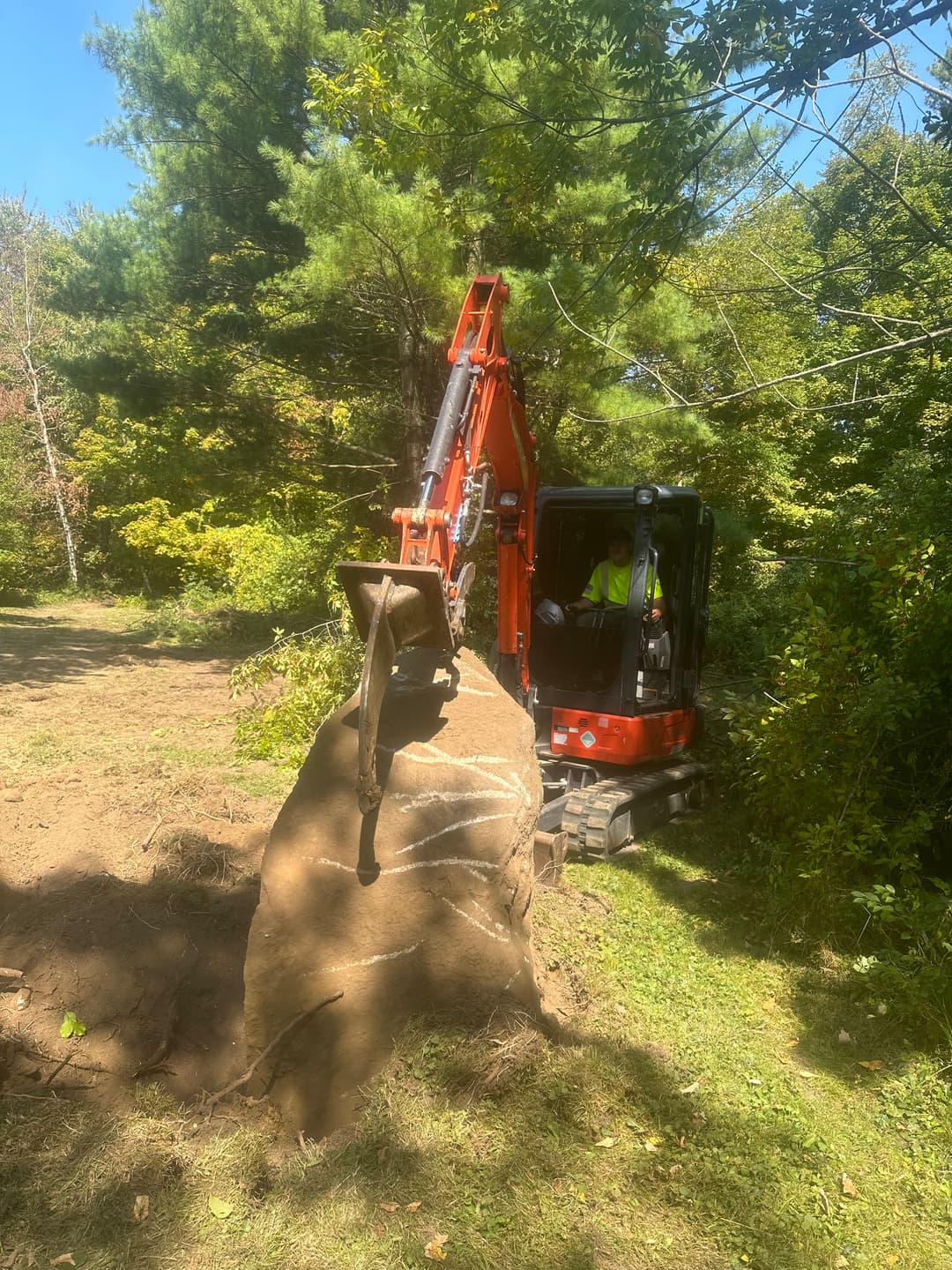 Excavator removing a large rock in a wooded area on a sunny day.